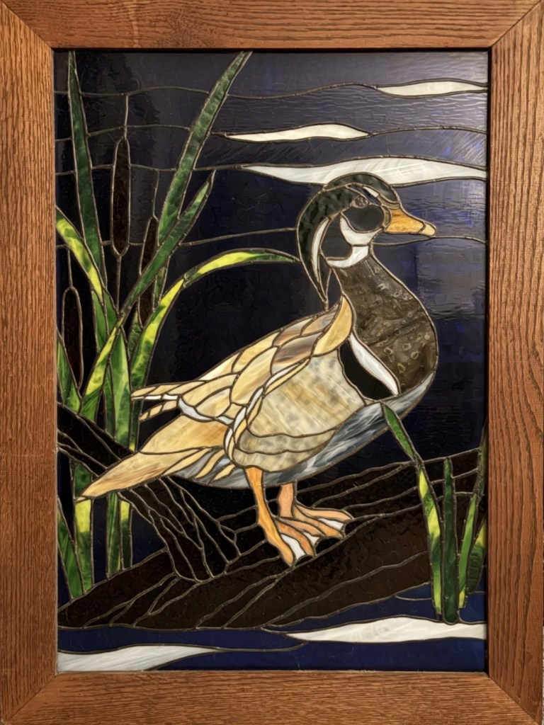 Framed Stained Glass piece of a male duck standing on a log in a pond surrounded by reeds.